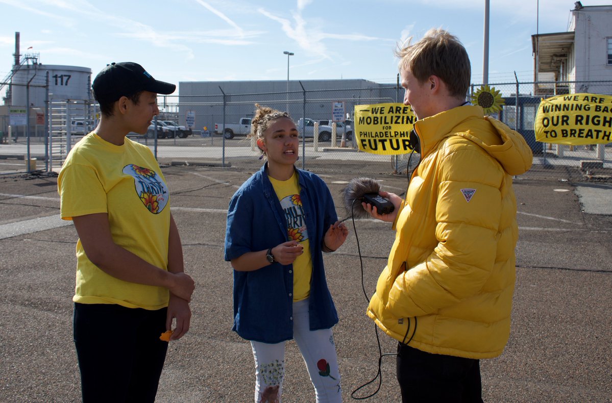This is a photo of a student in a bright yellow winter jacket holding a microphone, interviewing a pair also in yellow shirts.