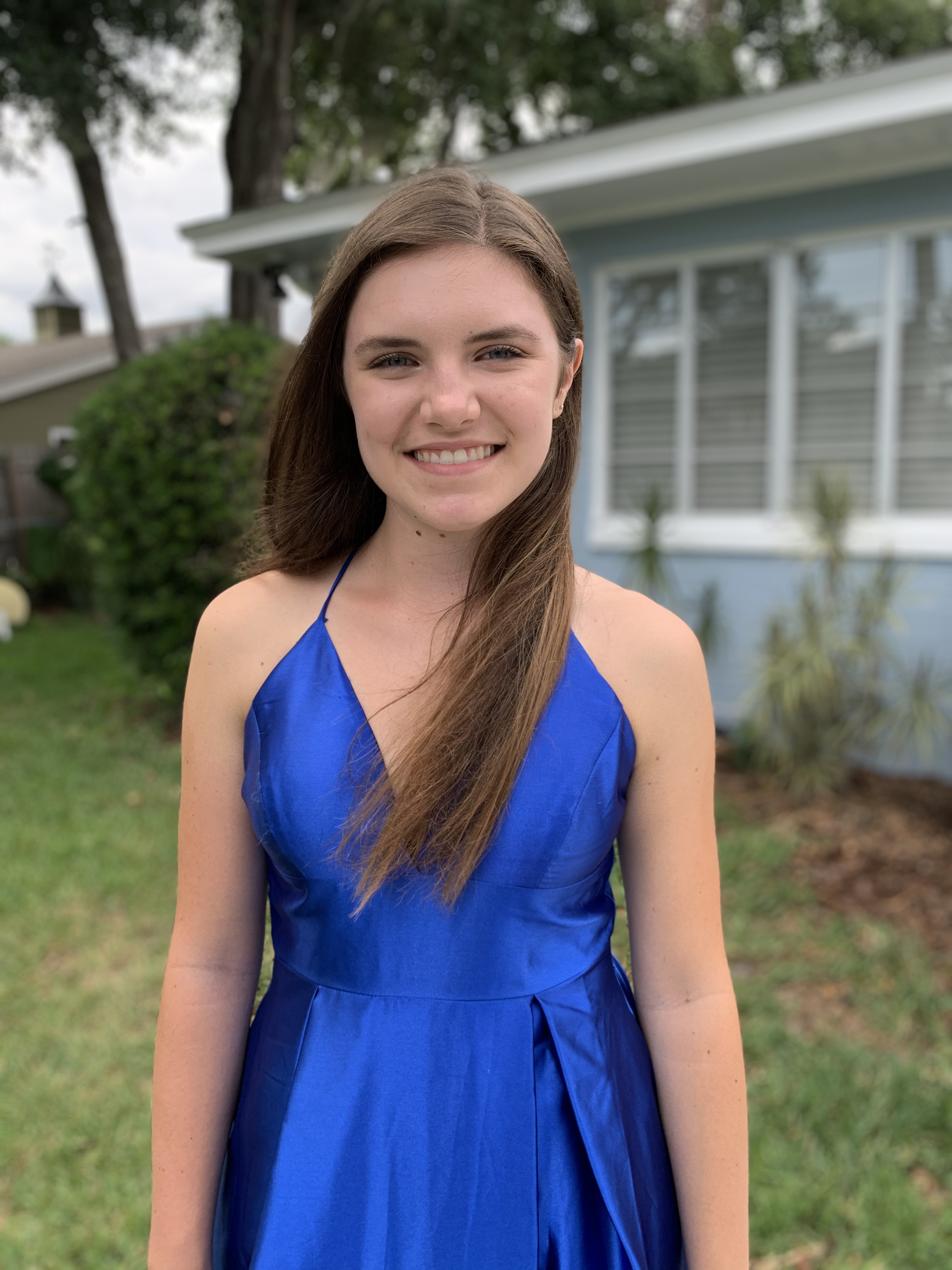 A person with long brown hair stands in front of a house. They are smiling and wearing a blue dress..