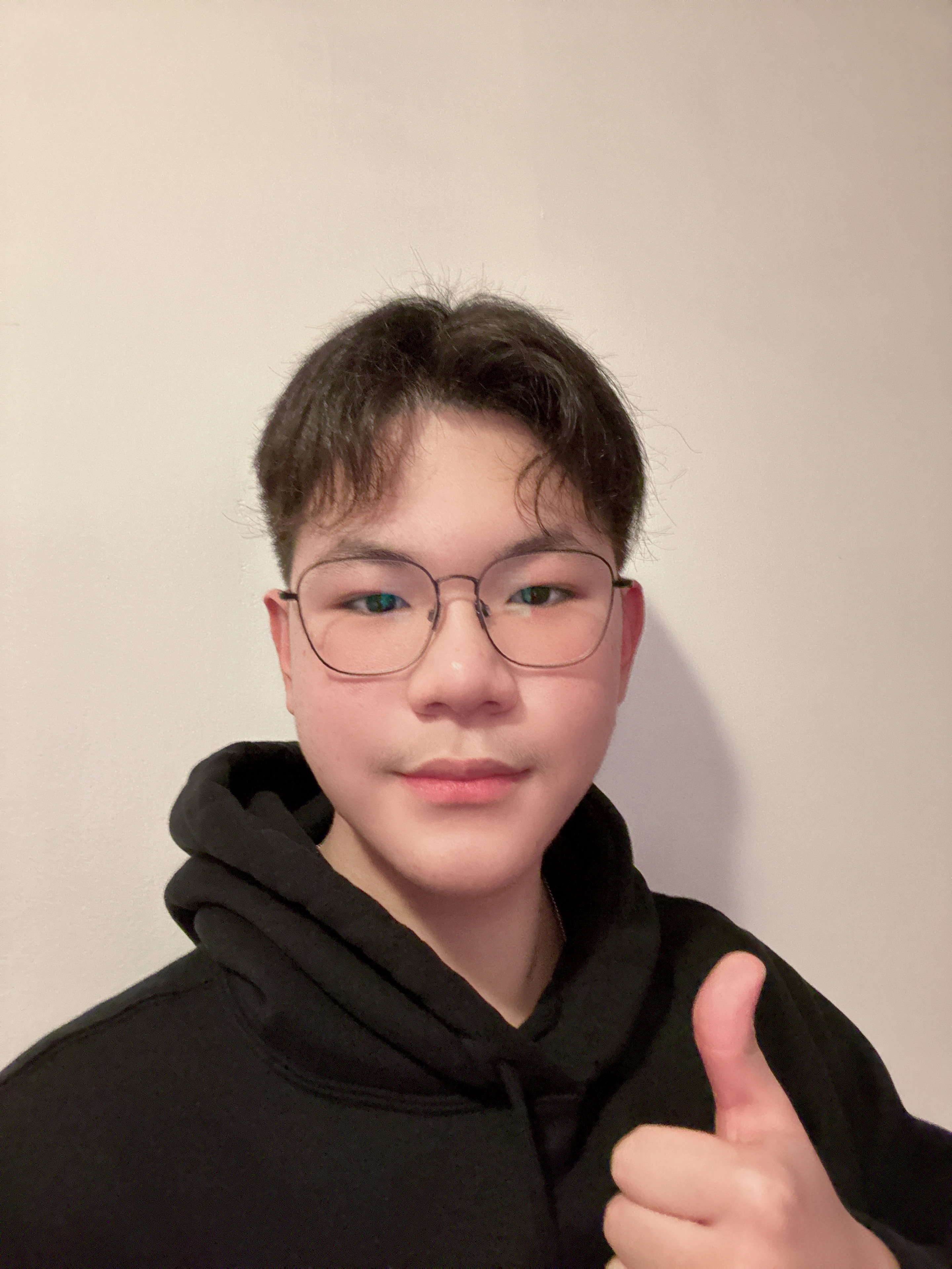 Yinle gives a thumbs up while wearing a black hoodie and black rimmed glasses. 