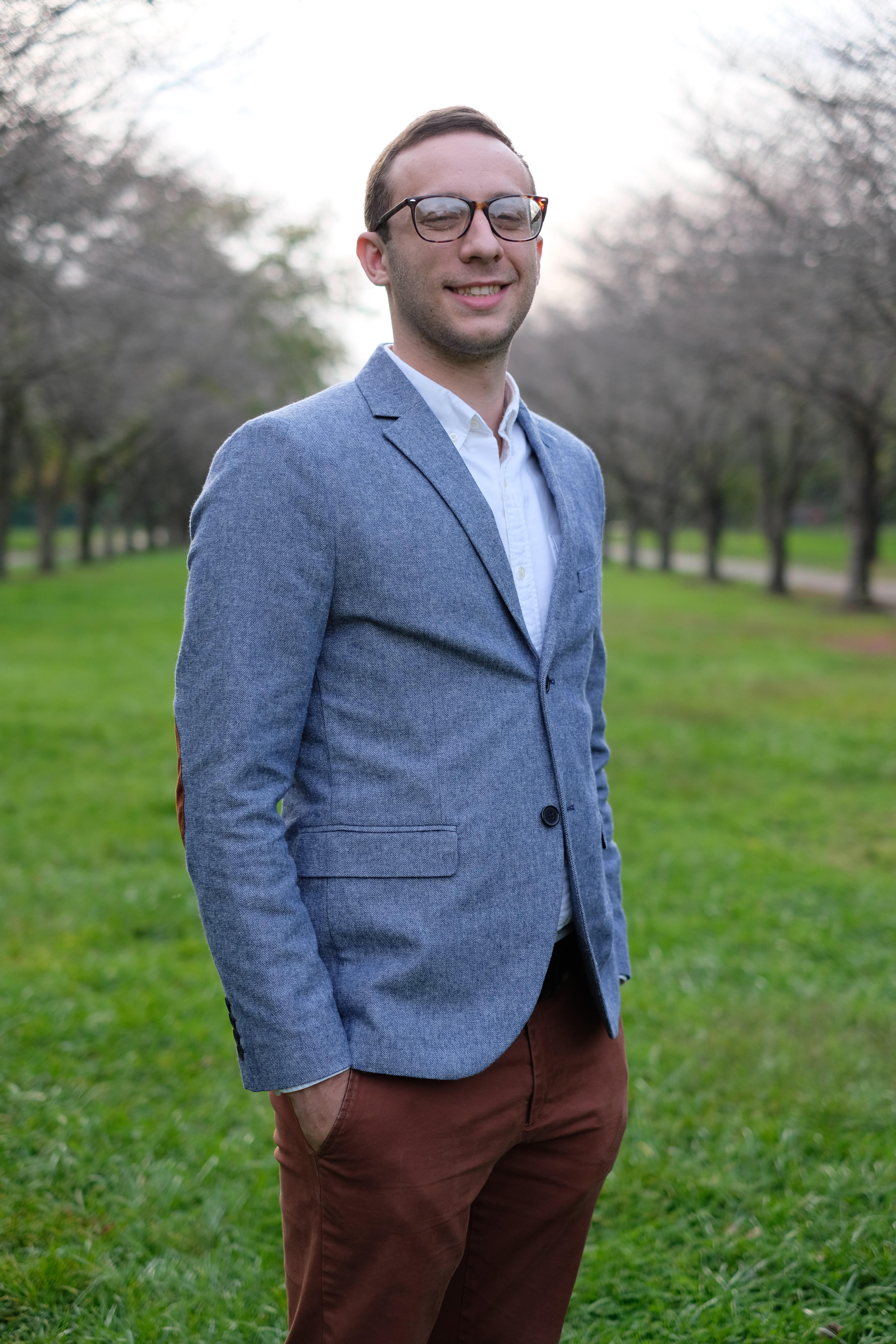 Man in a blue blazer and tortoiseshell glasses stands in a field and smiles at the camera.